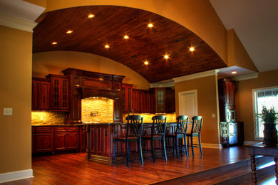 Large elegant u-shaped medium tone wood floor open concept kitchen photo in Other with an island, raised-panel cabinets, dark wood cabinets, granite countertops, gray backsplash, stone tile backsplash, stainless steel appliances and a farmhouse sink