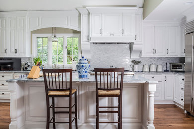 Example of a mid-sized classic l-shaped medium tone wood floor kitchen design in New York with raised-panel cabinets, white cabinets, marble countertops, white backsplash, stone tile backsplash, stainless steel appliances, an island and a farmhouse sink