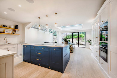 This is an example of a large classic l-shaped kitchen/diner in London with shaker cabinets, blue cabinets, white splashback, light hardwood flooring, an island, beige floors, white worktops, feature lighting, quartz worktops and engineered quartz splashback.