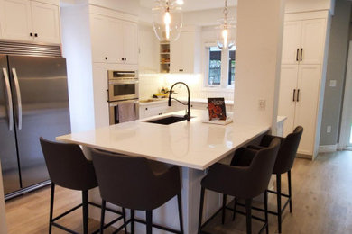 Large elegant l-shaped vinyl floor and brown floor kitchen photo in Toronto with a single-bowl sink, shaker cabinets, white cabinets, quartz countertops, white backsplash, ceramic backsplash, stainless steel appliances, an island and white countertops
