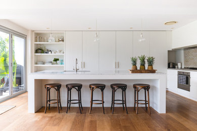 This is an example of a large contemporary l-shaped kitchen in Sydney with an undermount sink, flat-panel cabinets, white cabinets, mirror splashback, panelled appliances, with island, brown floor and white benchtop.