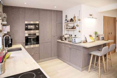 Example of a mid-sized trendy u-shaped light wood floor and white floor open concept kitchen design in London with an undermount sink, flat-panel cabinets, gray cabinets, wood countertops, white backsplash, window backsplash, paneled appliances and a peninsula