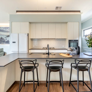 Photo of a contemporary galley open plan kitchen in Adelaide with an undermount sink, flat-panel cabinets, white cabinets, white splashback, mosaic tile splashback, white appliances, medium hardwood floors, with island, brown floor and grey benchtop.