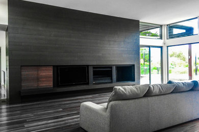 Modelo de sala de estar moderna con paredes grises, suelo de madera en tonos medios, estufa de leña, marco de chimenea de baldosas y/o azulejos, pared multimedia y suelo gris