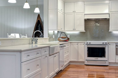 Design ideas for a mid-sized beach style l-shaped eat-in kitchen in Charleston with a farmhouse sink, shaker cabinets, white cabinets, quartz benchtops, grey splashback, glass tile splashback, white appliances, medium hardwood floors, no island, brown floor and white benchtop.