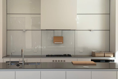 This is an example of a kitchen/diner in DC Metro with a single-bowl sink, flat-panel cabinets, white cabinets, engineered stone countertops, white splashback, glass sheet splashback, white appliances, light hardwood flooring, multiple islands and grey worktops.