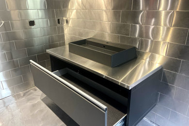Example of a trendy gray tile and metal tile concrete floor and gray floor powder room design in New York with flat-panel cabinets, black cabinets, a one-piece toilet, gray walls, a vessel sink, stainless steel countertops, gray countertops and a floating vanity