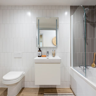 Immagine di una stanza da bagno minimalista con ante lisce, ante bianche, vasca da incasso, vasca/doccia, WC monopezzo, piastrelle grigie, piastrelle in ceramica, pareti bianche, pavimento in laminato, lavabo da incasso e porta doccia a battente