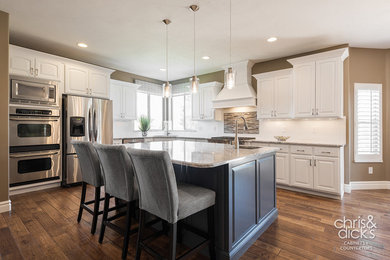Example of a large classic l-shaped medium tone wood floor open concept kitchen design in Salt Lake City with an undermount sink, raised-panel cabinets, white cabinets, quartz countertops, white backsplash, ceramic backsplash, stainless steel appliances and an island