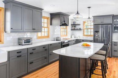 Inspiration for a large timeless l-shaped medium tone wood floor and brown floor eat-in kitchen remodel in Boston with a farmhouse sink, recessed-panel cabinets, gray cabinets, quartzite countertops, white backsplash, ceramic backsplash, stainless steel appliances, an island and white countertops