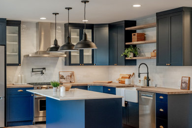 Example of a transitional l-shaped brown floor and medium tone wood floor open concept kitchen design in Seattle with shaker cabinets, blue cabinets, white backsplash, subway tile backsplash, stainless steel appliances, an island, white countertops, a farmhouse sink and quartz countertops