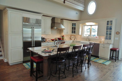 Mid-sized transitional l-shaped medium tone wood floor eat-in kitchen photo in New York with a farmhouse sink, raised-panel cabinets, white cabinets, granite countertops, beige backsplash, stainless steel appliances and an island