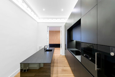Modelo de cocina lineal minimalista con armarios con paneles lisos, puertas de armario negras, electrodomésticos negros, fregadero de un seno, salpicadero negro, suelo de madera clara, una isla y encimeras negras