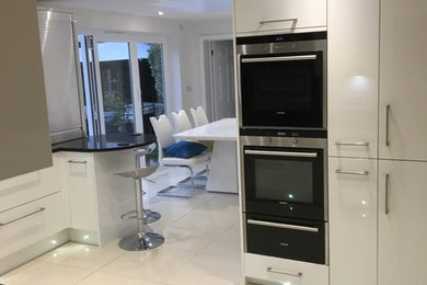 Medium sized contemporary l-shaped kitchen/diner in Hampshire with flat-panel cabinets, white cabinets, granite worktops, integrated appliances, ceramic flooring, a breakfast bar and white floors.