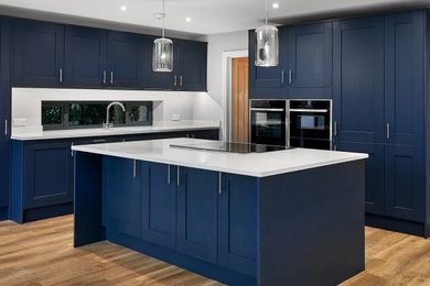Design ideas for a large traditional l-shaped open plan kitchen in Surrey with a built-in sink, shaker cabinets, blue cabinets, quartz worktops, black appliances, laminate floors, an island, brown floors and white worktops.