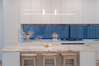 This is an example of an expansive modern l-shaped eat-in kitchen in Sydney with a single-bowl sink, white cabinets, marble benchtops, beige splashback, marble splashback, black appliances, ceramic floors, with island, beige floor and beige benchtop.