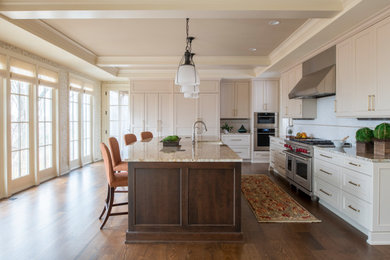 Large elegant l-shaped dark wood floor, brown floor and tray ceiling enclosed kitchen photo in Minneapolis with a farmhouse sink, recessed-panel cabinets, white cabinets, granite countertops, white backsplash, ceramic backsplash, stainless steel appliances, an island and multicolored countertops