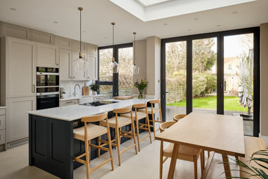 Photo of a medium sized traditional single-wall kitchen/diner in London with a belfast sink, shaker cabinets, grey cabinets, quartz worktops, grey splashback, engineered quartz splashback, stainless steel appliances, porcelain flooring, an island, beige floors and grey worktops.