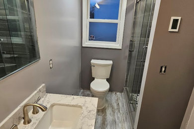 Bathroom - mid-sized 3/4 gray tile and porcelain tile laminate floor, brown floor and single-sink bathroom idea in New York with shaker cabinets, gray cabinets, a two-piece toilet, brown walls, an integrated sink, marble countertops, multicolored countertops and a freestanding vanity