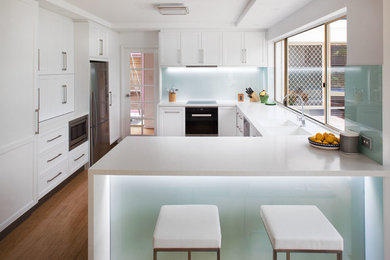 This is an example of a mid-sized transitional u-shaped kitchen in Perth with an integrated sink, recessed-panel cabinets, white cabinets, solid surface benchtops, white splashback, glass sheet splashback, stainless steel appliances, bamboo floors and a peninsula.