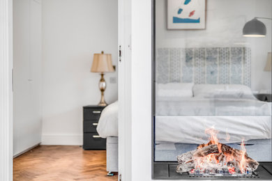 Imagen de dormitorio principal minimalista pequeño con paredes blancas, suelo de madera clara, chimenea de doble cara, marco de chimenea de metal y suelo marrón