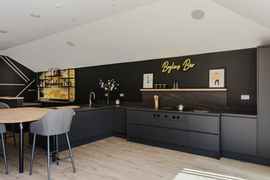 This is an example of a medium sized contemporary l-shaped open plan kitchen in Other with a submerged sink, flat-panel cabinets, black cabinets, black splashback, light hardwood flooring, no island, brown floors, black worktops and a feature wall.
