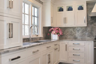 Transitional medium tone wood floor kitchen photo in New York with a drop-in sink, recessed-panel cabinets, white cabinets, glass countertops, gray backsplash, stone slab backsplash, paneled appliances and an island