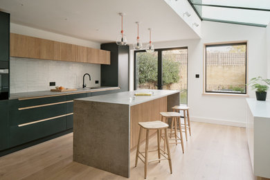 Large modern grey and white single-wall open plan kitchen in London with an integrated sink, painted wood flooring, an island, brown floors, grey worktops, black cabinets, white splashback and black appliances.