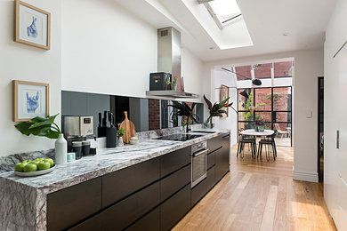 Photo of a mid-sized modern galley open plan kitchen with flat-panel cabinets, white cabinets, marble benchtops, black splashback, glass sheet splashback, stainless steel appliances, medium hardwood floors, no island, brown floor, white benchtop and an undermount sink.