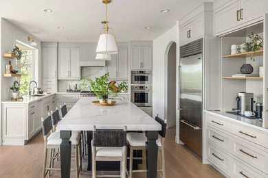 Mid-sized transitional l-shaped light wood floor and brown floor open concept kitchen photo in Seattle with an undermount sink, shaker cabinets, white cabinets, quartz countertops, white backsplash, quartz backsplash, stainless steel appliances, an island and white countertops