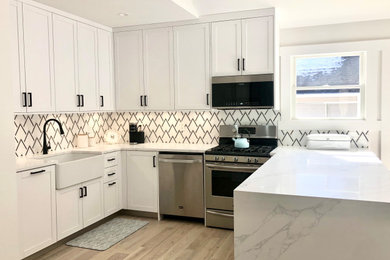 Example of a mid-sized trendy u-shaped light wood floor, brown floor and vaulted ceiling kitchen pantry design in New York with a farmhouse sink, recessed-panel cabinets, white cabinets, marble countertops, multicolored backsplash, ceramic backsplash, stainless steel appliances, no island and white countertops