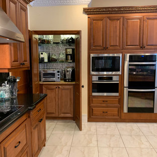 Cette photo montre une très grande arrière-cuisine chic avec un évier 3 bacs, un placard avec porte à panneau surélevé, des portes de placard en bois brun, un plan de travail en granite, une crédence noire, une crédence en dalle de pierre, un électroménager en acier inoxydable, un sol en carrelage de porcelaine, un îlot central, un sol beige et un plan de travail noir.