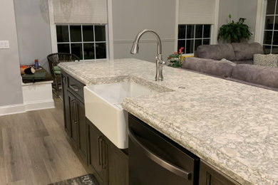 Photo of a mid-sized transitional l-shaped open plan kitchen in Cleveland with a farmhouse sink, flat-panel cabinets, white cabinets, quartz benchtops, white splashback, porcelain splashback, black appliances, medium hardwood floors, with island, beige floor, multi-coloured benchtop and vaulted.