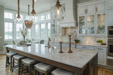 Beach style medium tone wood floor and brown floor eat-in kitchen photo in Tampa with a farmhouse sink, raised-panel cabinets, white cabinets, beige backsplash, mosaic tile backsplash, stainless steel appliances, an island and beige countertops