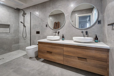 This is an example of a medium sized modern ensuite wet room bathroom in Melbourne with flat-panel cabinets, medium wood cabinets, grey tiles, porcelain tiles, grey walls, porcelain flooring, a vessel sink, engineered stone worktops, grey floors, an open shower, white worktops, a wall niche, double sinks and a floating vanity unit.