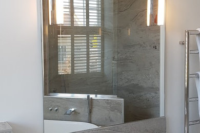 This is an example of a large contemporary kids wet room bathroom in Other with flat-panel cabinets, white cabinets, a freestanding tub, a wall-mount toilet, gray tile, porcelain tile, grey walls, porcelain floors, a wall-mount sink, marble benchtops, grey floor, an open shower and white benchtops.