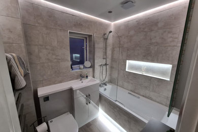 Photo of a medium sized contemporary family bathroom in Cheshire with flat-panel cabinets, grey cabinets, a built-in bath, a shower/bath combination, a two-piece toilet, beige tiles, stone tiles, beige walls, laminate floors, a built-in sink, solid surface worktops, beige floors, a hinged door, white worktops, a single sink and a built in vanity unit.