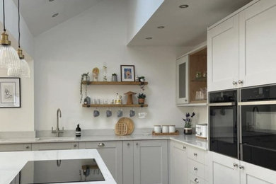 Exemple d'une grande cuisine ouverte chic avec un placard avec porte à panneau encastré, des portes de placard blanches, un plan de travail en quartz modifié, un électroménager noir, parquet clair, îlot, un plan de travail blanc et un plafond voûté.