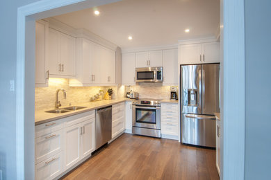 Small modern l-shaped separate kitchen in Toronto with a double-bowl sink, shaker cabinets, white cabinets, solid surface benchtops, grey splashback, stone tile splashback, stainless steel appliances, medium hardwood floors, no island and brown floor.