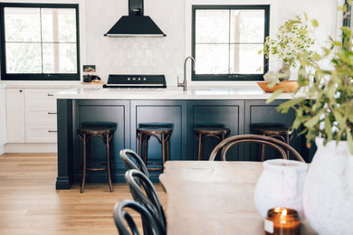 Inspiration for a mid-sized country l-shaped eat-in kitchen in Gold Coast - Tweed with an undermount sink, shaker cabinets, black cabinets, quartz benchtops, white splashback, subway tile splashback, black appliances, light hardwood floors, with island and white benchtop.