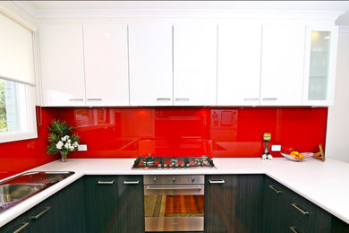This is an example of a mid-sized contemporary u-shaped eat-in kitchen in Melbourne with a double-bowl sink, flat-panel cabinets, white cabinets, laminate benchtops, red splashback, glass sheet splashback, stainless steel appliances and no island.