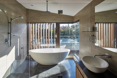 This is an example of a contemporary master wet room bathroom in Sydney with flat-panel cabinets, medium wood cabinets, a freestanding tub, brown tile, brown walls, a vessel sink, black floor, a hinged shower door and black benchtops.