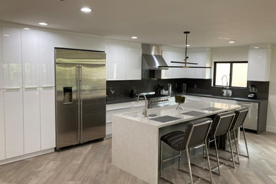 Example of a l-shaped light wood floor kitchen design in Los Angeles with an undermount sink, flat-panel cabinets, white cabinets, quartz countertops, stainless steel appliances, an island, black backsplash, quartz backsplash and black countertops