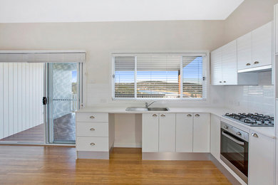 Mid-sized contemporary l-shaped open plan kitchen in Central Coast with a single-bowl sink, louvered cabinets, white cabinets, laminate benchtops, white splashback, ceramic splashback, stainless steel appliances, medium hardwood floors, no island and beige floor.