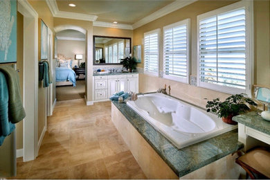 Inspiration for a large coastal master beige tile and limestone tile limestone floor, beige floor, double-sink and vaulted ceiling bathroom remodel in Orange County with raised-panel cabinets, white cabinets, a one-piece toilet, beige walls, a drop-in sink, granite countertops, green countertops and a built-in vanity