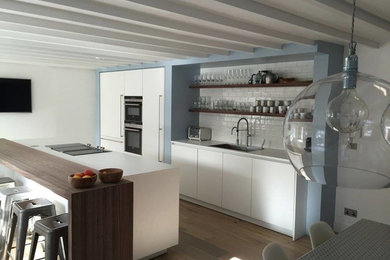 Photo of a single-wall open plan kitchen in Edinburgh with a single-bowl sink, flat-panel cabinets, white cabinets, white splashback, stainless steel appliances, medium hardwood floors, with island and white benchtop.