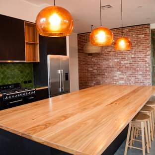 Exemple d'une arrière-cuisine tendance en L avec un évier 2 bacs, un placard à porte plane, des portes de placard noires, un plan de travail en bois, une crédence verte, une crédence en carreau de terre cuite, un électroménager en acier inoxydable, béton au sol, un îlot central et un sol gris.