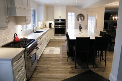 Mid-sized trendy l-shaped medium tone wood floor and brown floor eat-in kitchen photo in Toronto with a double-bowl sink, recessed-panel cabinets, white cabinets, white backsplash, subway tile backsplash, stainless steel appliances, an island, quartz countertops and white countertops