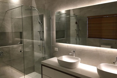 This is an example of a mid-sized contemporary master bathroom in Melbourne with flat-panel cabinets, a double shower, gray tile, porcelain tile, a trough sink, engineered quartz benchtops, a hinged shower door and white benchtops.