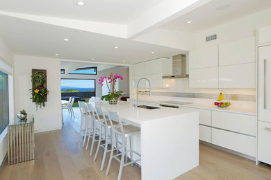 Photo of a mid-sized modern single-wall open plan kitchen in Los Angeles with an undermount sink, flat-panel cabinets, white cabinets, solid surface benchtops, stainless steel appliances, light hardwood floors, with island, metallic splashback and glass tile splashback.